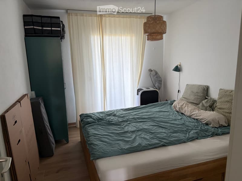 Cozy bedroom with a wooden bed, blue duvet, pillows, a wardrobe, a hanging lamp, and a fan on a table.