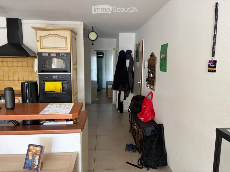 Hallway with tiled floor, coat rack, and a small table with a picture frame and papers. Kitchen area visible with oven, countertop, and a speaker.