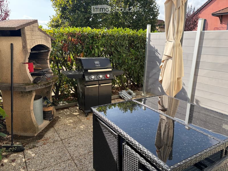 Outdoor patio area with a stone pizza oven, a modern grill, a wicker table and chairs, and a large umbrella. The space is enclosed by a white fence and surrounded by greenery.