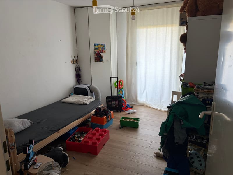 A small, well-lit bedroom with a single bed, a small stool, a fan, and various toys and storage boxes scattered around. The room features wooden flooring, white walls, and a large window with curtains.