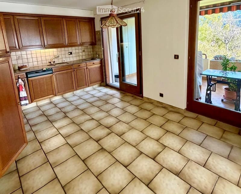 Kitchen with wooden cabinets, tiled floor, sink, and oven. Sliding glass door leads to a balcony with a view and a small table with a potted plant.