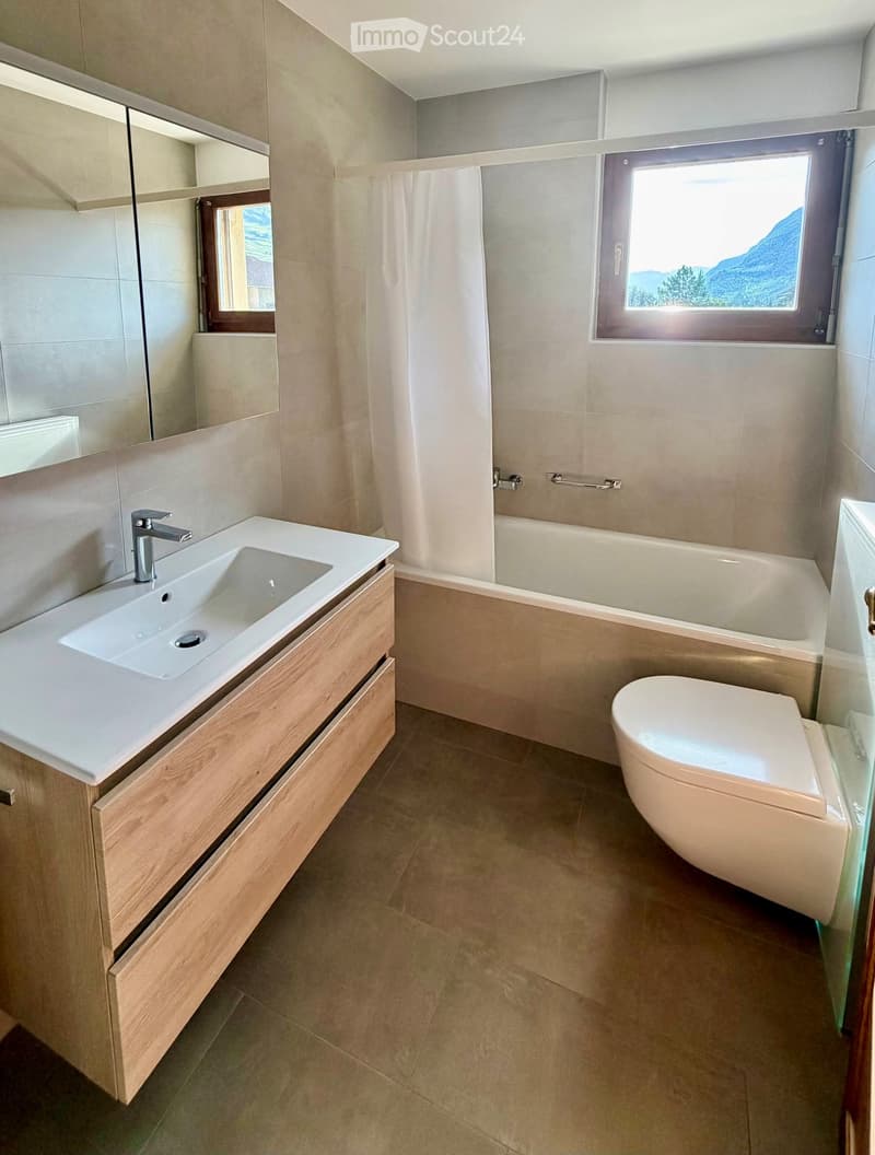 Modern bathroom with a white sink, wooden cabinet, bathtub, toilet, and large windows offering a scenic mountain view.