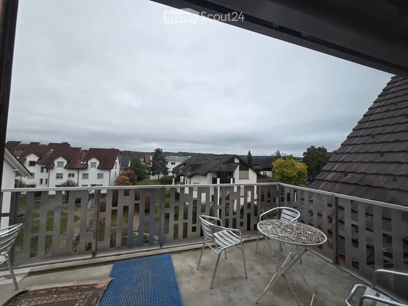 Balcony with metal railing, small round table, and chairs, overlooking a residential area with houses and trees.