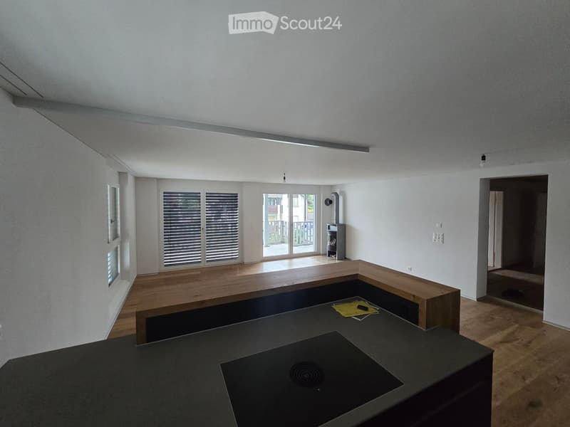 Modern, minimalist room with a dark countertop, wooden bench seating, and a small fireplace. Large windows and sliding glass doors lead to a balcony area.