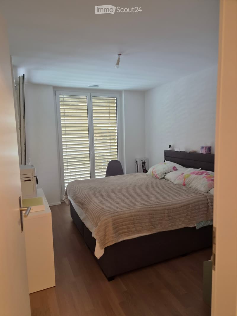 A modern bedroom with a large bed, a nightstand, a chair, and a mirror. The room features wooden flooring and large windows with blinds.