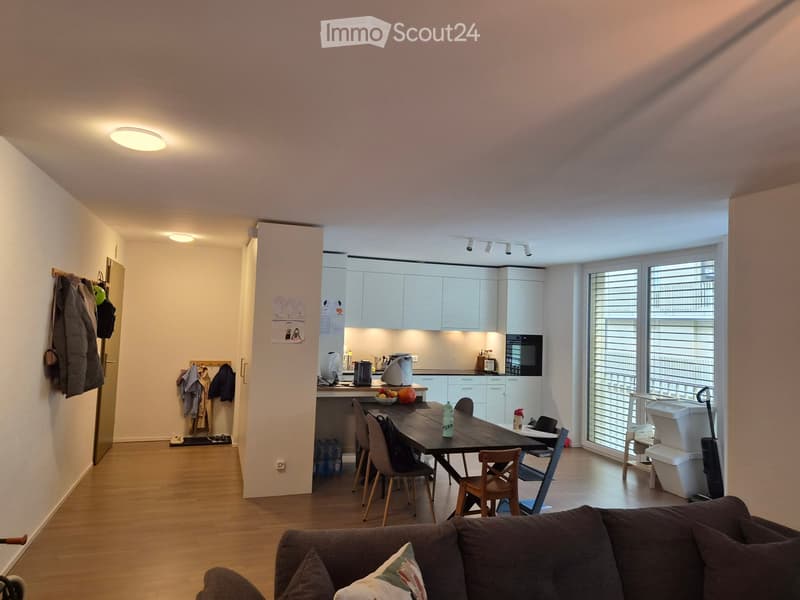 Open-plan living and dining area with a sofa, dining table, chairs, and a modern kitchen featuring white cabinets, an oven, and a refrigerator. The space includes a coat rack and a coat hanging on the wall.