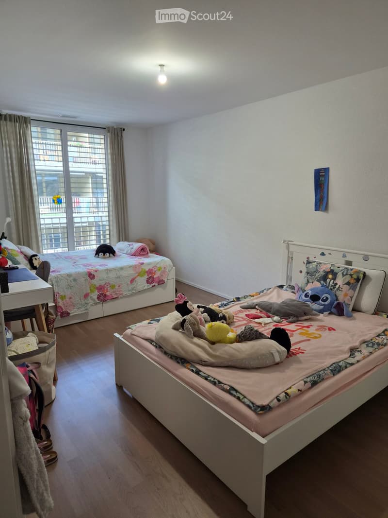 A well-lit bedroom with two single beds, each with colorful bedding and stuffed toys, a small stool, and a handbag near the door.