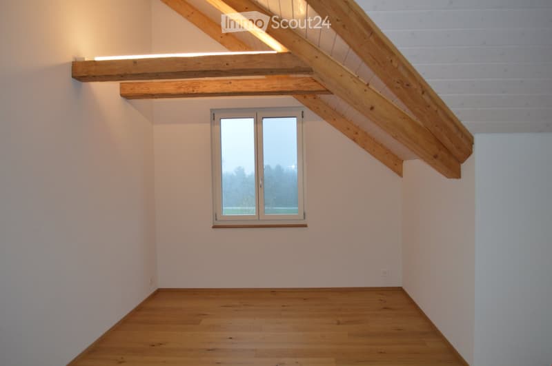 Empty attic room with wooden beams, white walls, wooden floor, and a window offering a view of the outdoors.