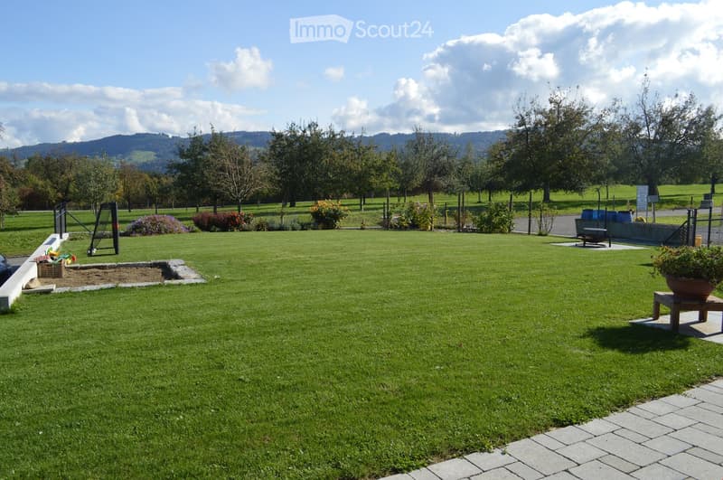 Well-maintained lawn with a small sandbox, a few potted plants, and a bench, surrounded by trees and a fence with a scenic mountain backdrop.