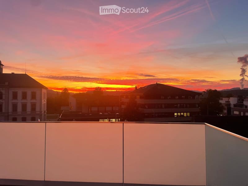 View from a rooftop terrace at sunset, showing cityscape with buildings and distant hills.