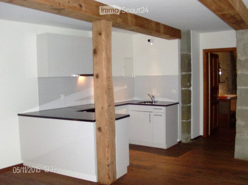 Modern kitchen with white cabinets, black countertops, and a central island. Wooden floor and exposed wooden beam in the center.