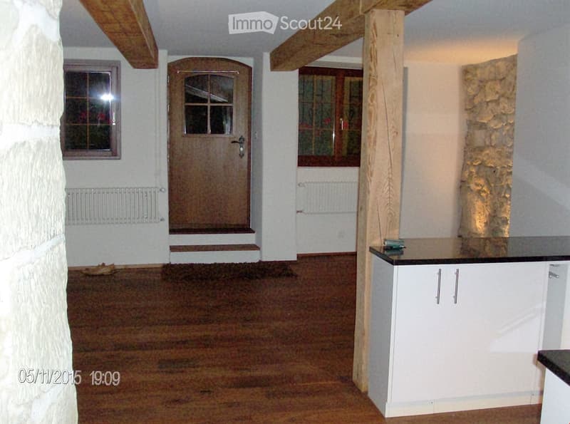 A partially furnished room with a wooden floor, white walls, a stone pillar, a cabinet with a countertop, and a wooden door with glass panels.