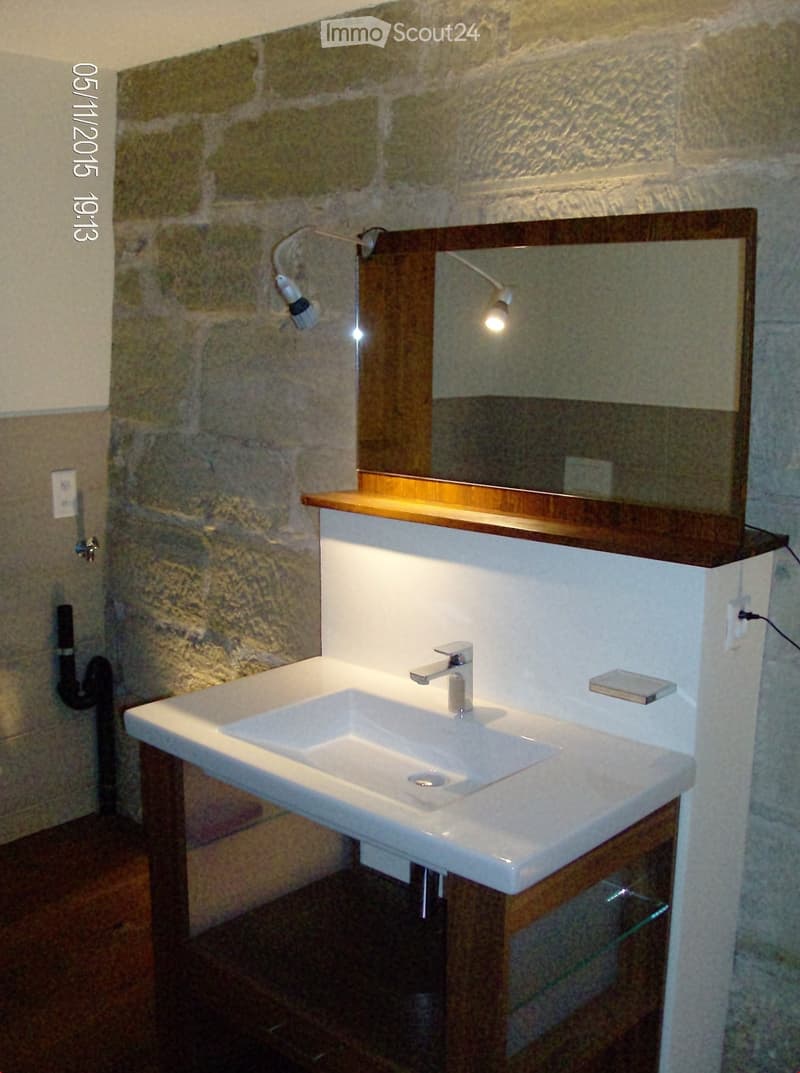 Modern bathroom with a white sink, chrome faucet, wooden shelf, and large mirror above it. The walls are made of stone, and the floor is tiled.
