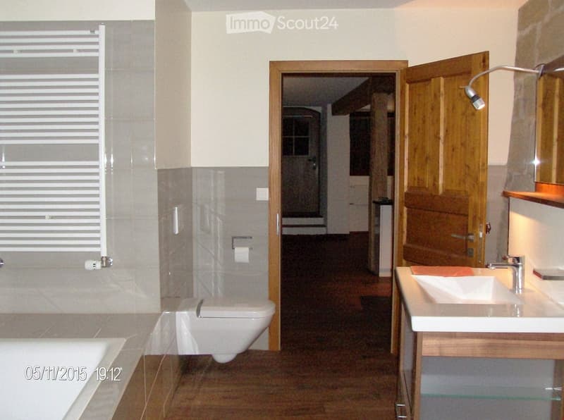 Modern bathroom with a toilet, sink, bathtub, and mirror. The room features tiled walls and a wooden door leading to another area.