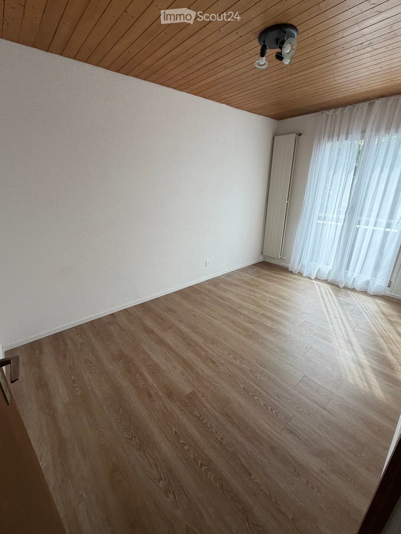 Empty room with wooden flooring, white walls, and a wooden ceiling. A window with sheer curtains is visible on one wall, and a radiator is present below the window.