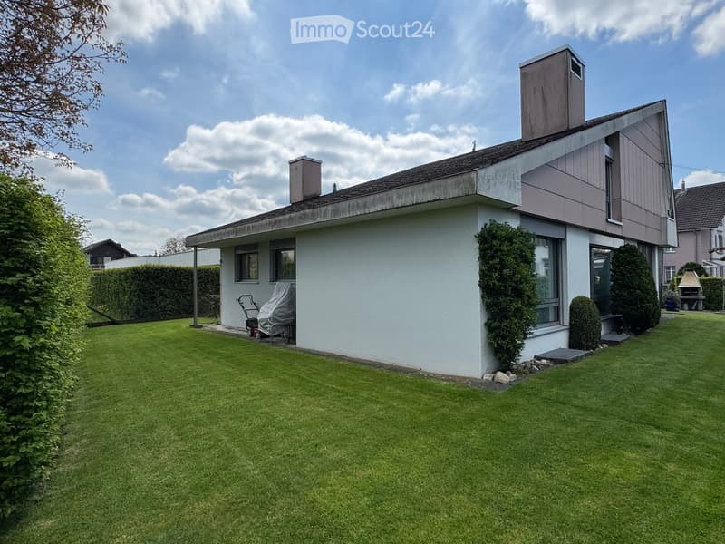 Well-maintained lawn with trimmed hedges and a modern house in the background, featuring large windows and a covered patio area.
