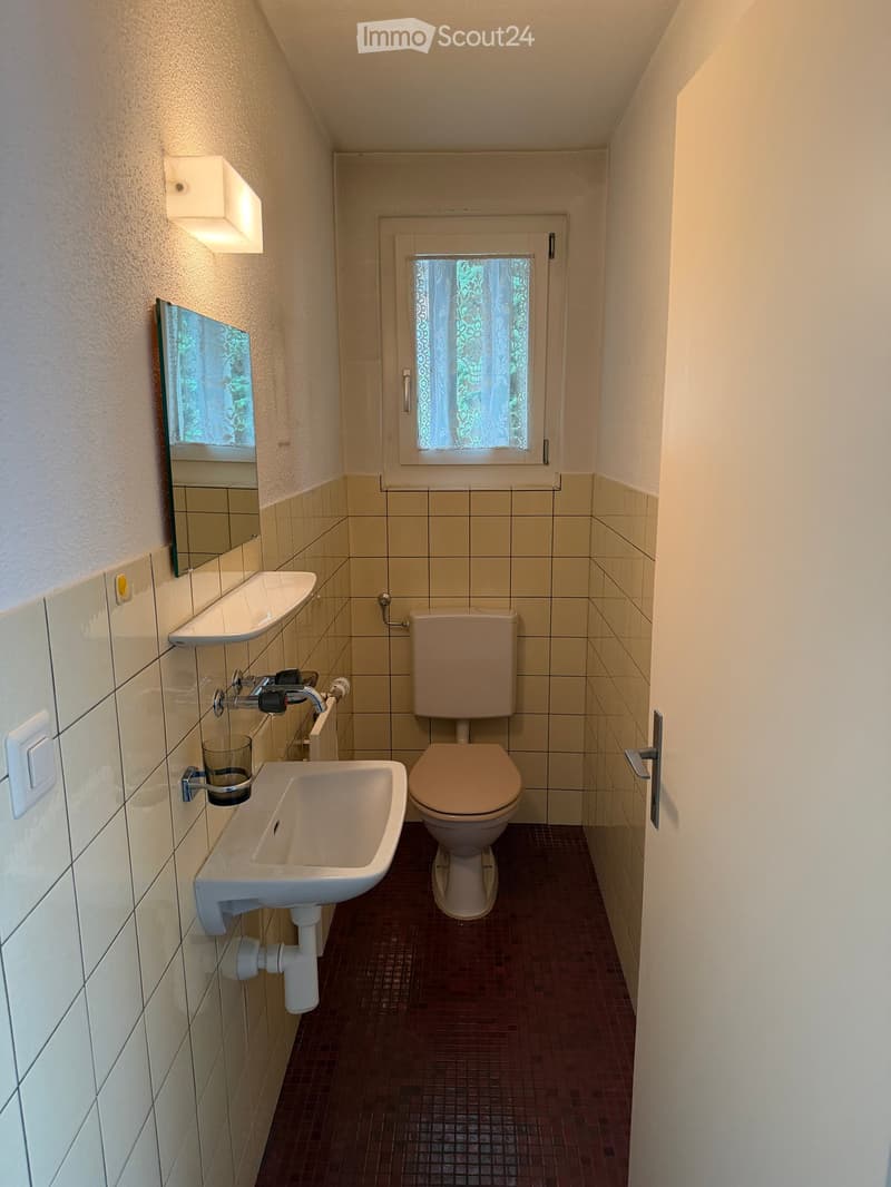 Small bathroom with beige tiles, a toilet, a sink, a mirror, and a window with a curtain.