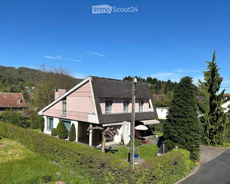 Two-story house with a sloped roof, pink siding, and white trim. Features a balcony, patio area with outdoor furniture, and is surrounded by greenery and trees.