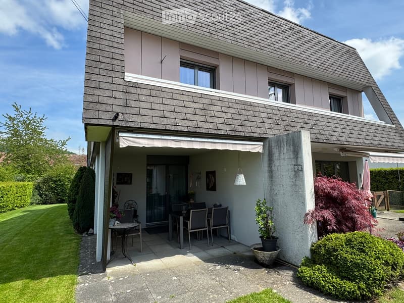 Two-story house with a modern design, featuring a covered patio area with a dining table and chairs, surrounded by a well-maintained garden and greenery.