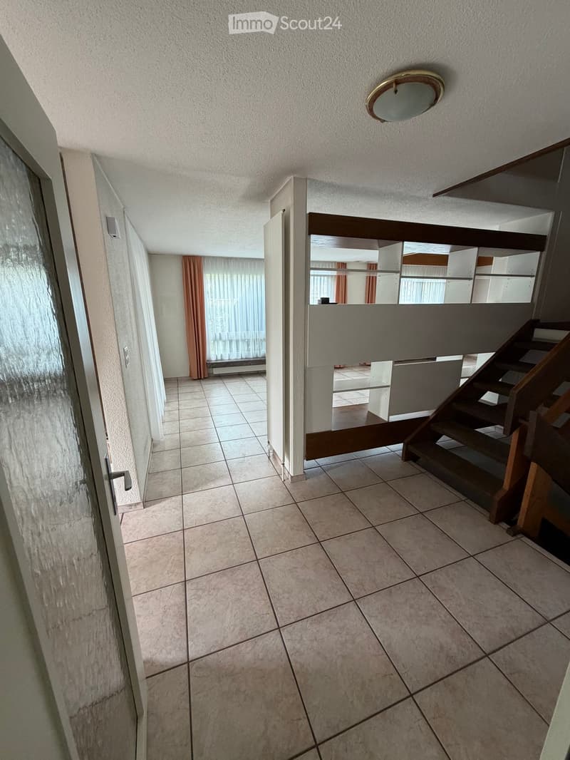 Empty hallway with tiled floor, wooden staircase, and open doorway leading to another room.