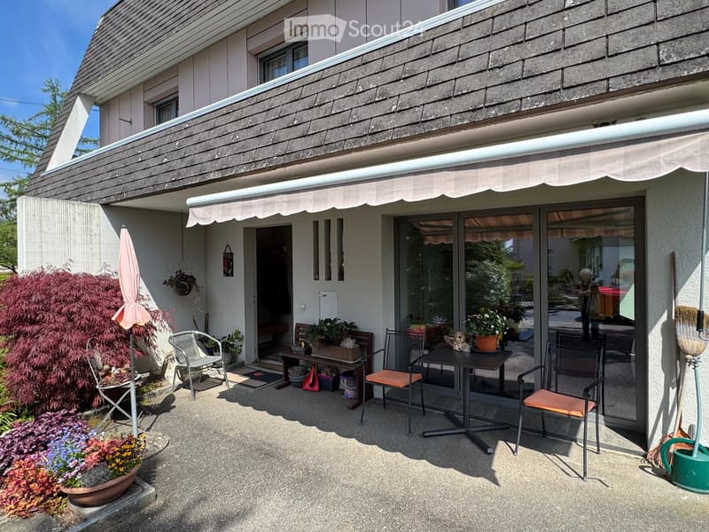 Balcony with a table, chairs, umbrella, and flower pots, attached to a house with a covered awning and glass doors.