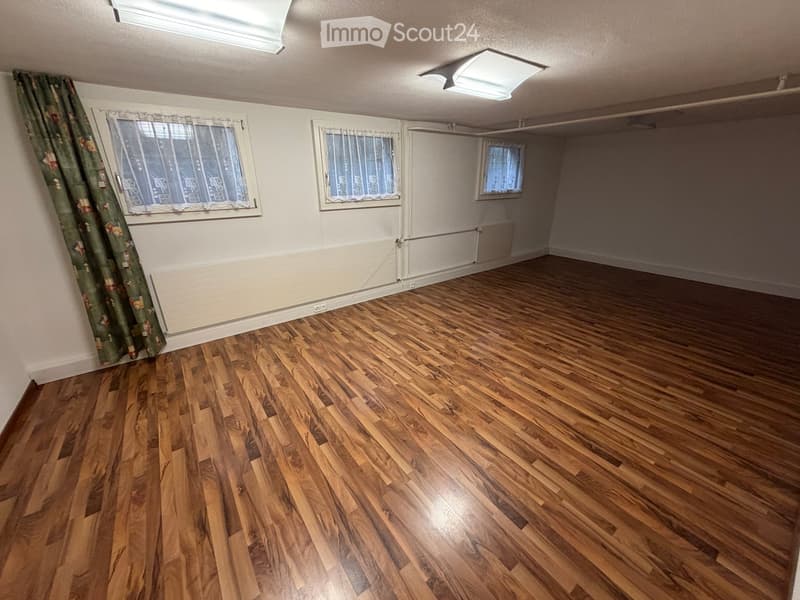 Empty room with wooden flooring, white walls, and three windows with curtains. The room has a radiator under each window and a ceiling light fixture.