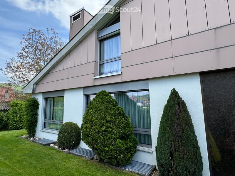 Modern two-story house with light-colored exterior, large windows, and well-maintained garden with trimmed bushes.