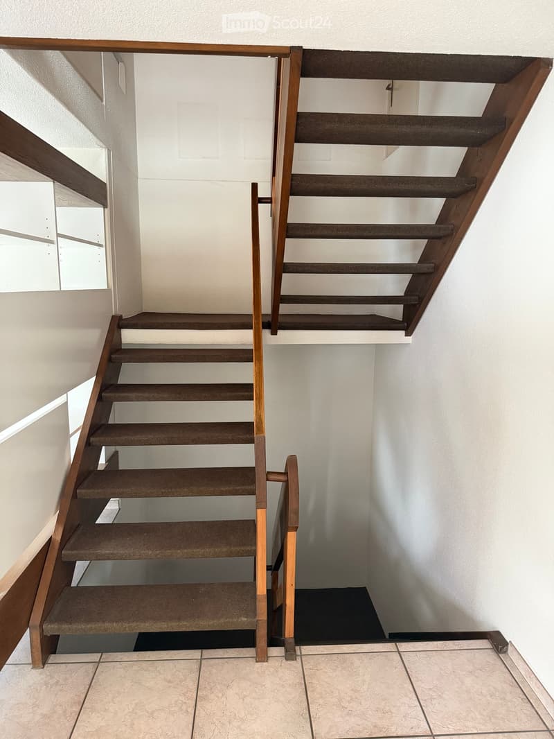 Modern staircase with wooden handrails and carpeted steps, leading up to a higher level with white walls and tiled flooring at the base.