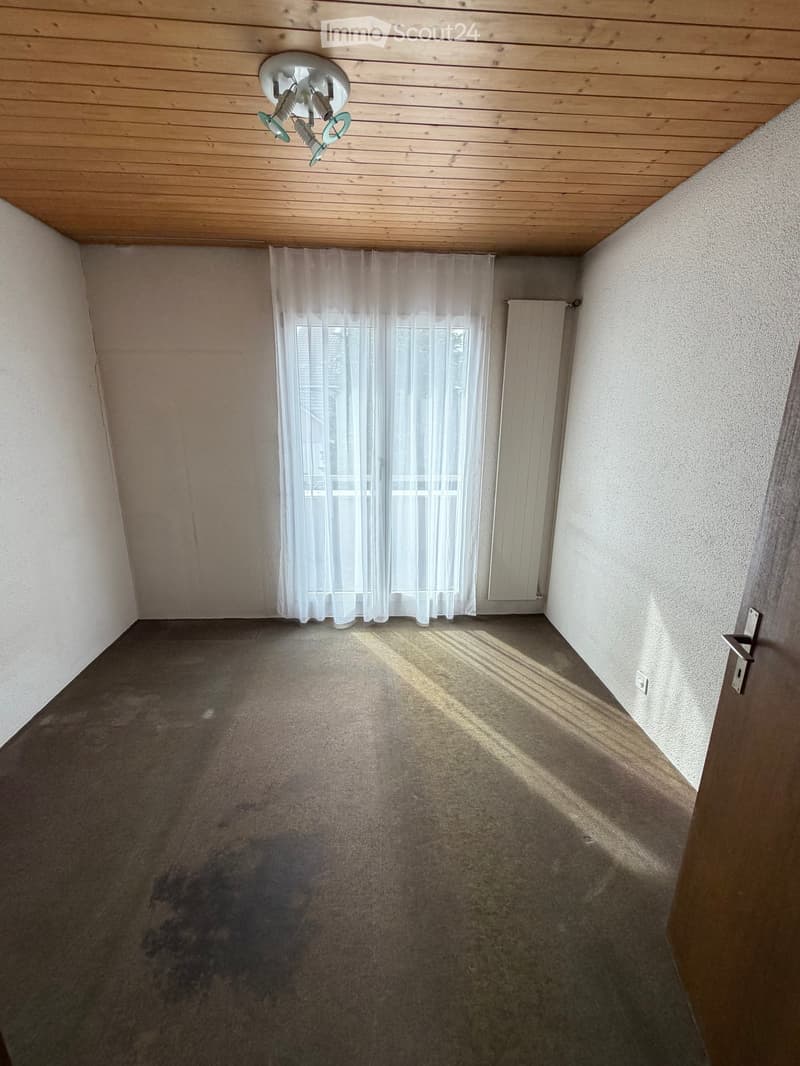 Empty room with light-colored walls, carpeted floor, and a window with white curtains. A radiator is visible on one wall, and a ceiling light fixture is present.
