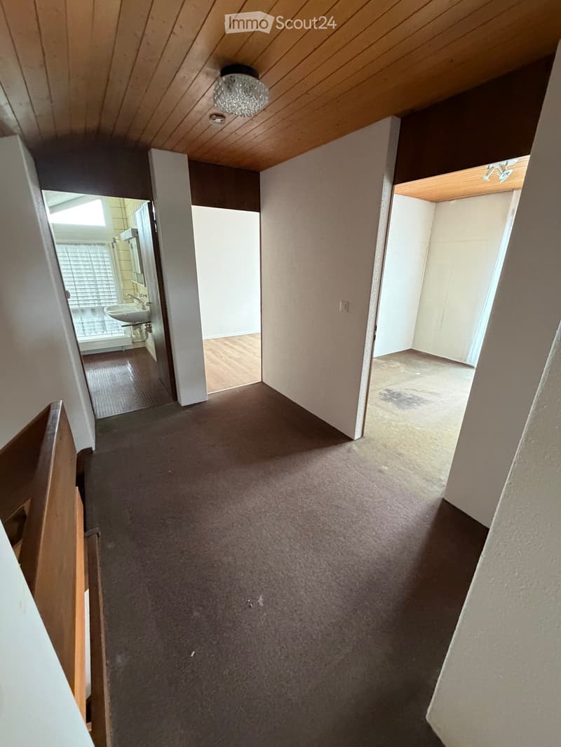 Carpeted hallway with wooden ceiling, leading to a bathroom and another room. The bathroom has a sink and a window with blinds.