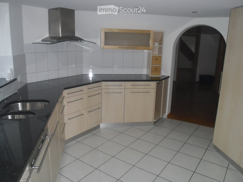 Modern kitchen with light wooden cabinets, black countertops, stainless steel sink, and a range hood. The floor is tiled, and there is a small microwave on the counter.