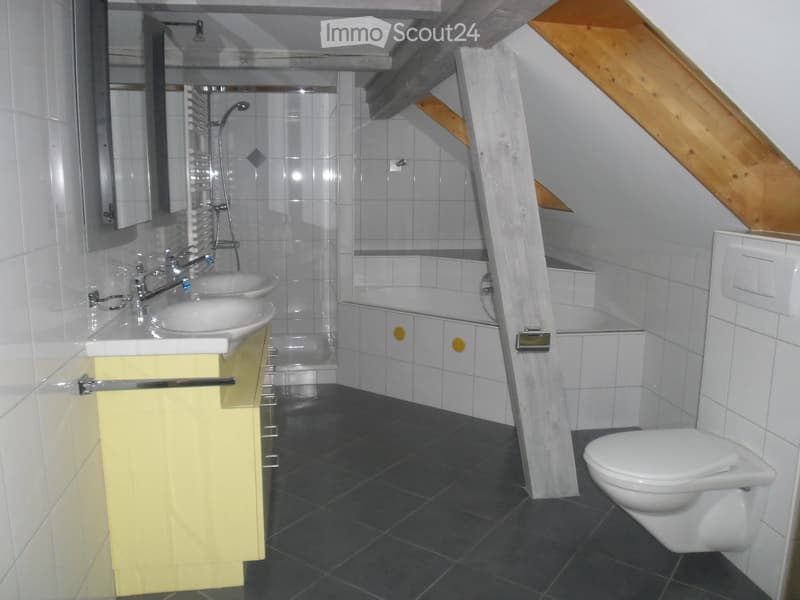 Modern bathroom with double sinks, a bathtub, a toilet, and a mirror. The space features white tiles and a sloped wooden ceiling.