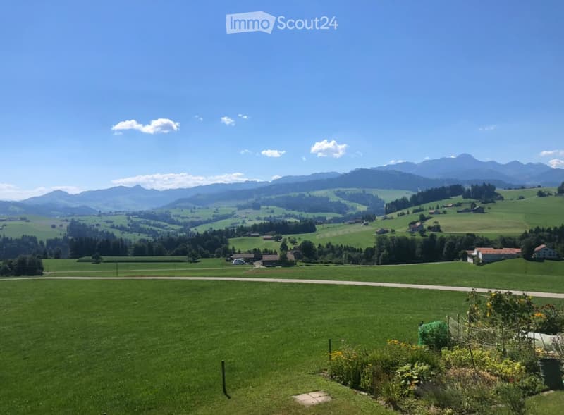 A panoramic view of a lush green valley with rolling hills, scattered houses, and distant mountains under a clear blue sky.