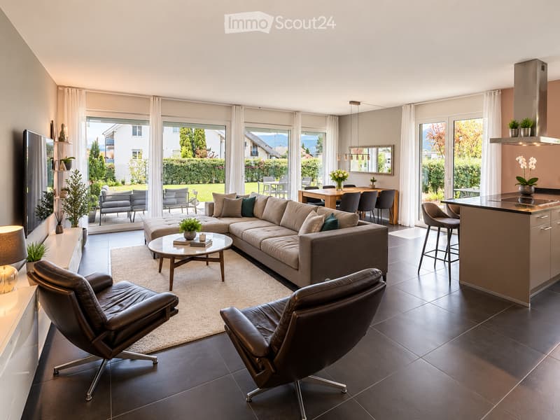 Modern open-plan living and dining area with large windows offering a view of the garden. Features a grey sectional sofa, coffee table, leather chairs, kitchen island, dining table, and decorative items.