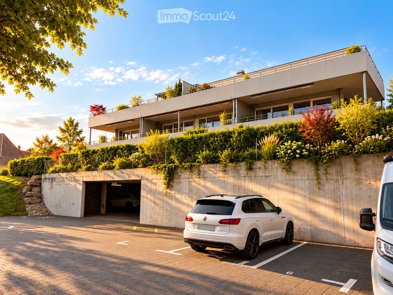 Modern two-story building with a rooftop terrace, surrounded by greenery, with a covered garage entrance and a parking area.