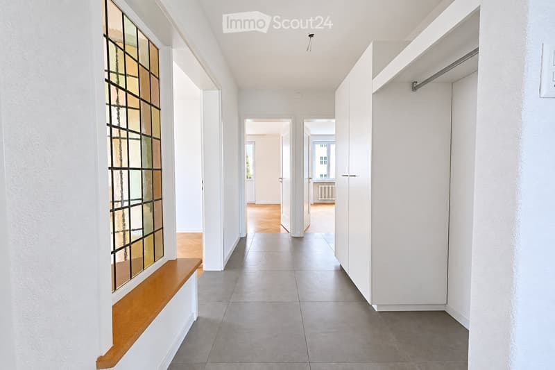 Modern hallway with white walls, grey tiled floor, wooden bench, and stained glass window. Open doors lead to adjacent rooms with wooden flooring.