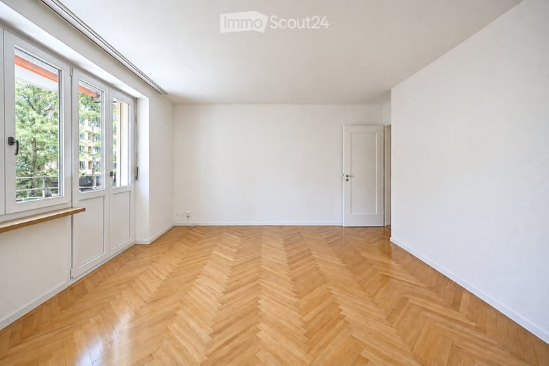 Empty room with light wooden herringbone flooring, white walls, large windows with white frames, and a white door leading to another space.