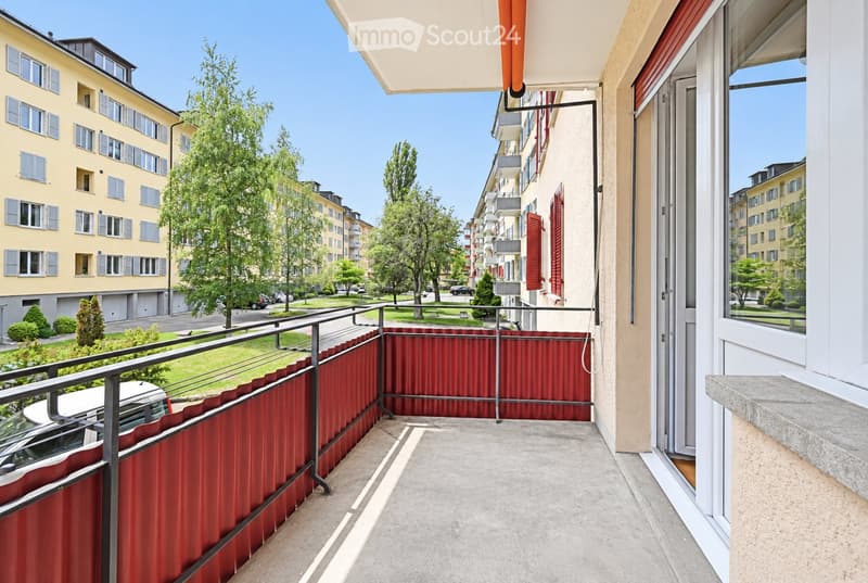 Balcony with metal railing, red privacy screen, and view of a residential street with trees and buildings.