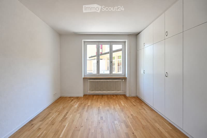 Empty room with light wooden flooring, white walls, large window with radiator below, and built-in white closet units.