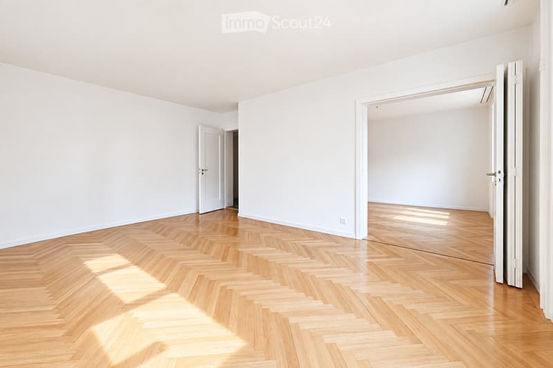 Empty room with white walls and herringbone wooden flooring, featuring two open doors leading to adjacent spaces.