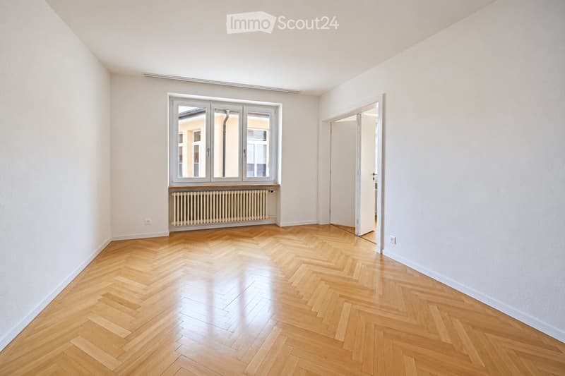 Empty room with light-colored walls, herringbone wooden flooring, large window with white frame, radiator below window, and an open doorway leading to another space.