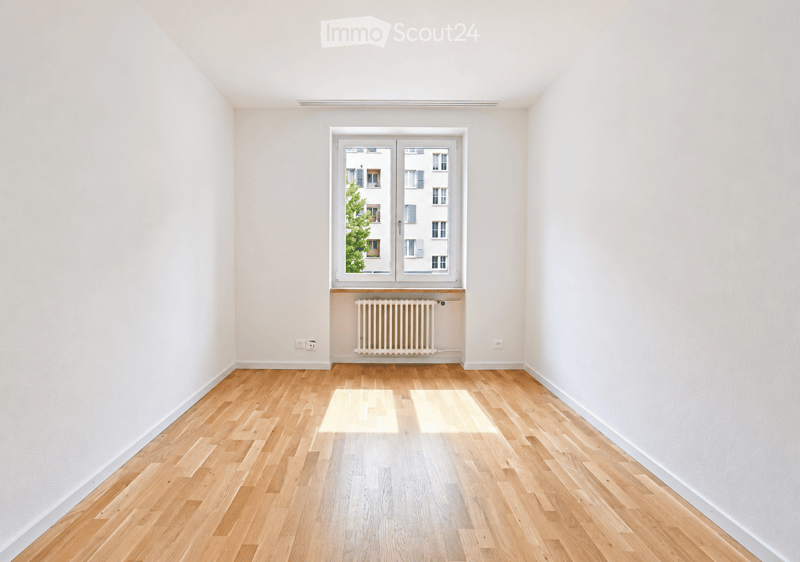 Empty room with light wooden flooring, white walls, and a large window with double doors leading to a view of a building and greenery outside. A radiator is visible below the window.