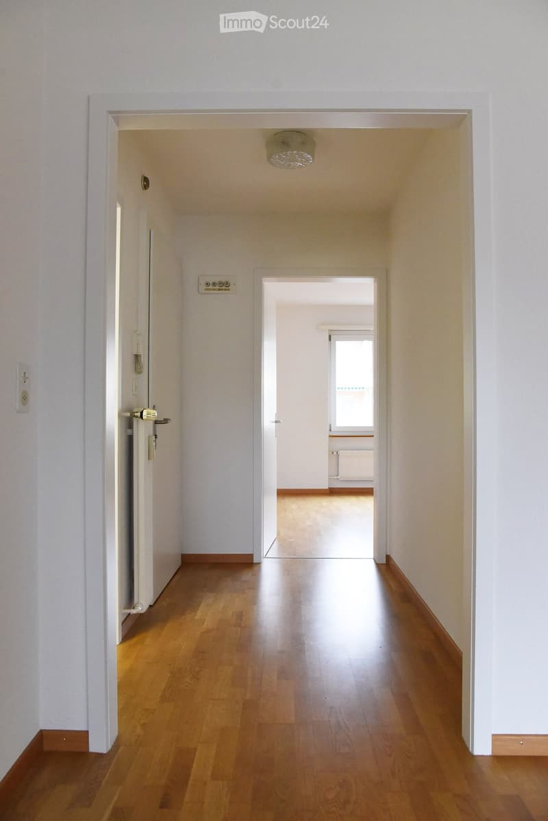 Empty hallway with wooden flooring, white walls, and a ceiling light fixture. Doors lead to adjacent rooms, one of which has a visible window and radiator.
