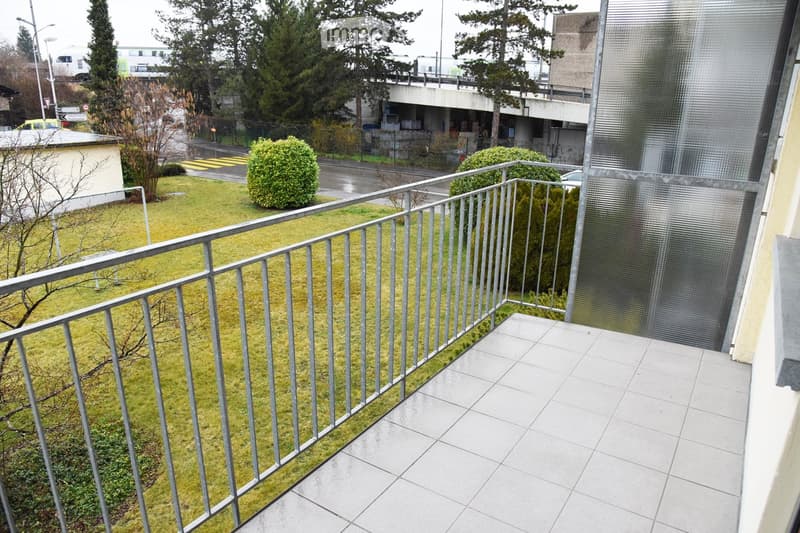 Balcony with tiled floor and metal railing, overlooking a grassy area with bushes and trees, with a road and buildings in the background.