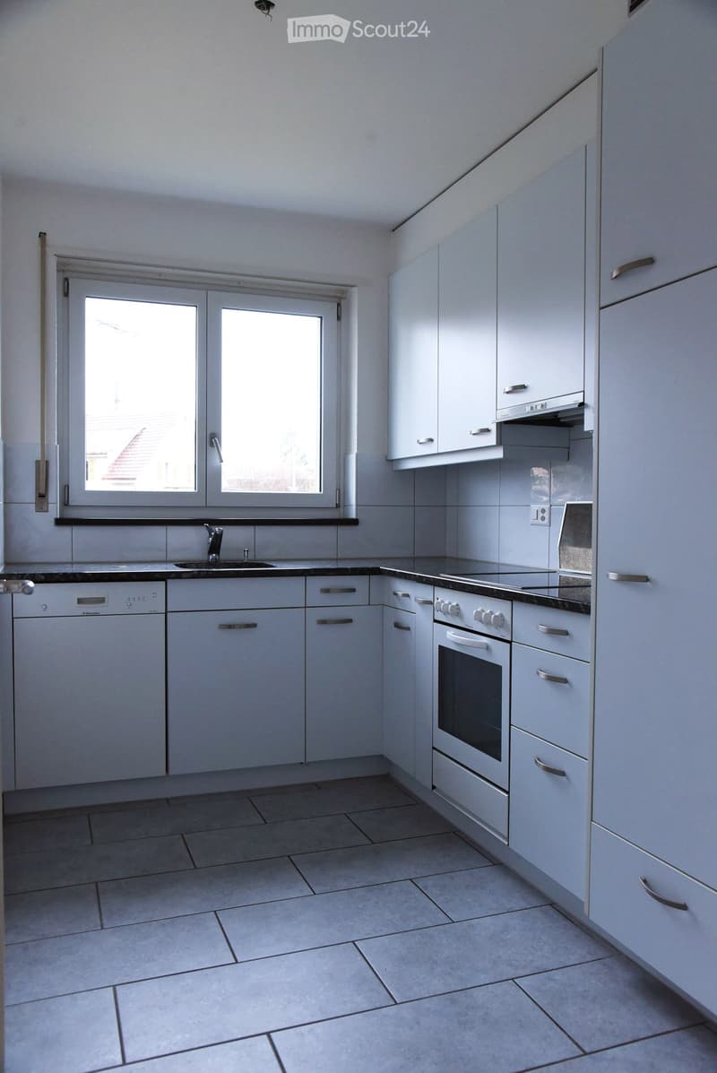 Modern kitchen with white cabinets, black countertops, a sink, an oven, and a dishwasher. The space is well-lit with large windows and tiled flooring.
