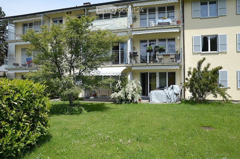 A multi-story residential building with balconies, awnings, and a well-maintained garden area with a covered patio and outdoor furniture.