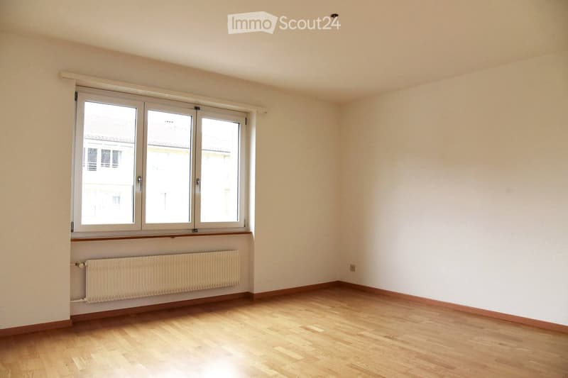 Empty room with wooden flooring, white walls, large windows, and a radiator below the windows.