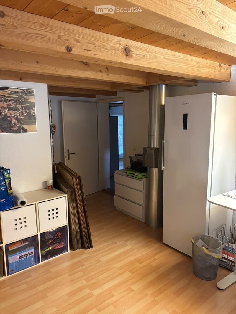 A small, well-lit room with wooden flooring and exposed wooden beams on the ceiling. It features a refrigerator, a storage unit with various items, a small table, and a trash bin. A door leads to another room, and a picture frame is mounted on the wall.
