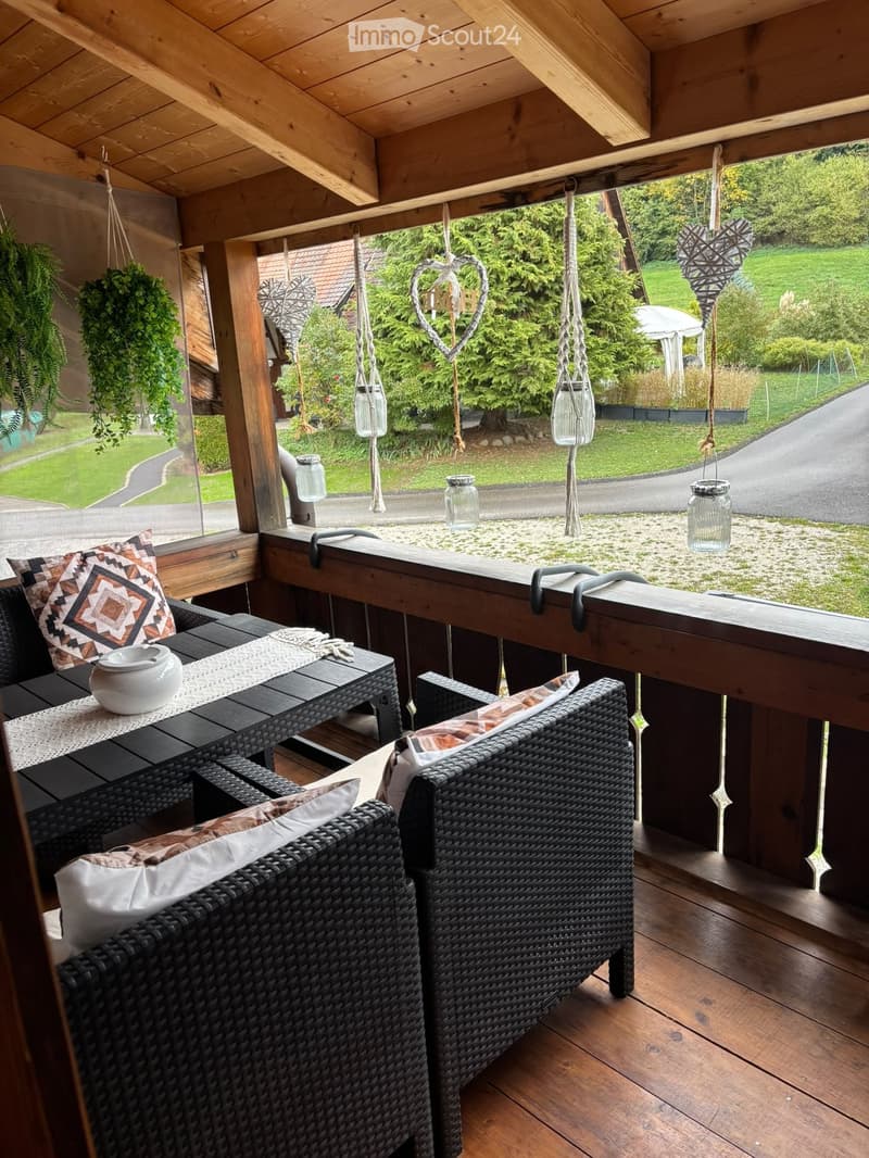 A wooden balcony with wicker furniture, including a sofa, chairs, and a small table with a vase, overlooking a green garden.