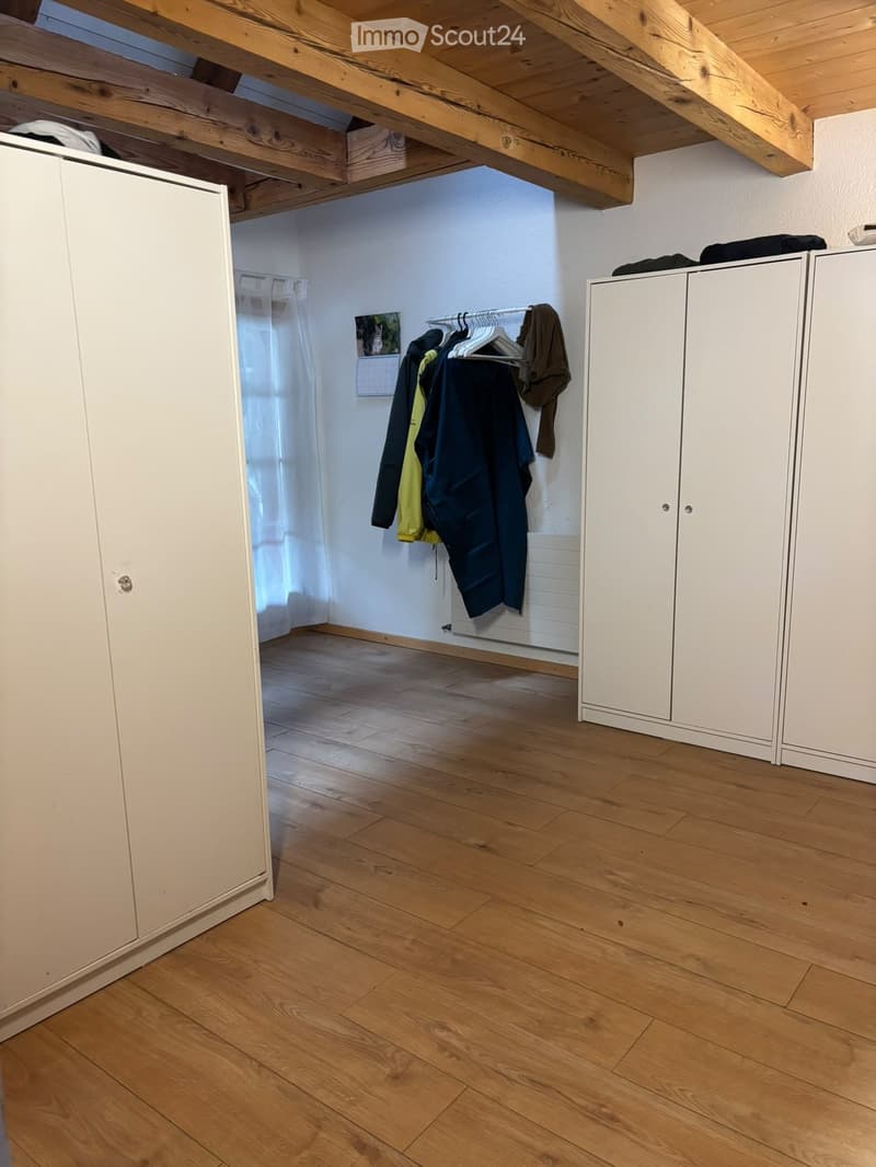 Empty room with wooden flooring, white built-in wardrobes, exposed wooden beams on the ceiling, and a coat rack with jackets and a calendar on the wall.